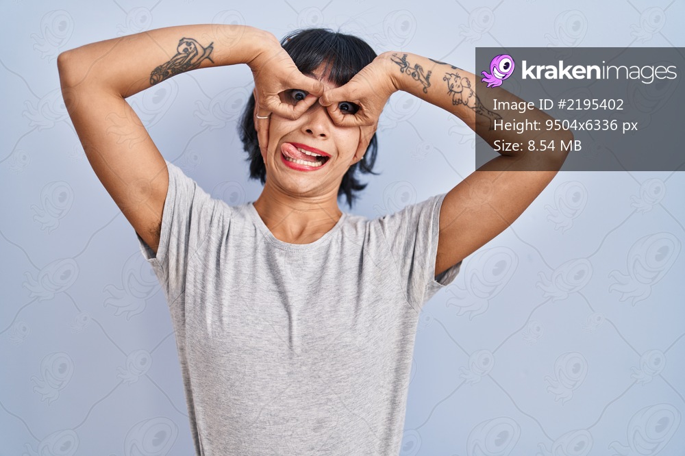Young hispanic woman wearing casual t shirt over blue background doing ok gesture like binoculars sticking tongue out, eyes looking through fingers. crazy expression. 