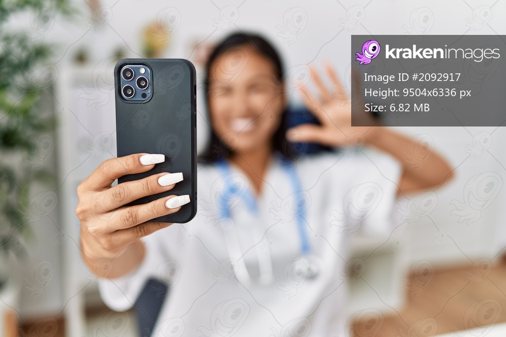 Young latin woman wearing doctor uniform make selfie by the smartphone at clinic