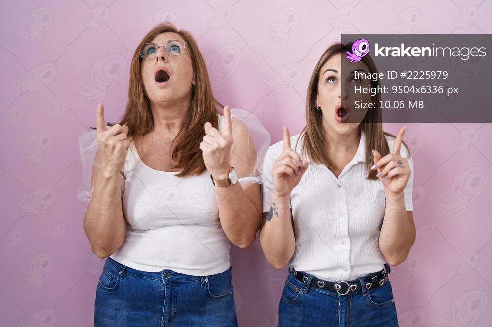 Hispanic mother and daughter wearing casual white t shirt over pink background amazed and surprised looking up and pointing with fingers and raised arms. 
