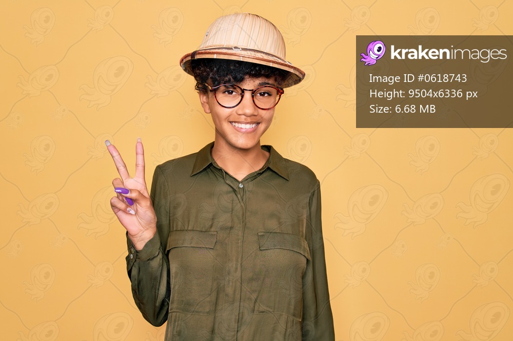 Young beautiful african american tourist woman wearing glasses and explorer hat showing and pointing up with fingers number two while smiling confident and happy.