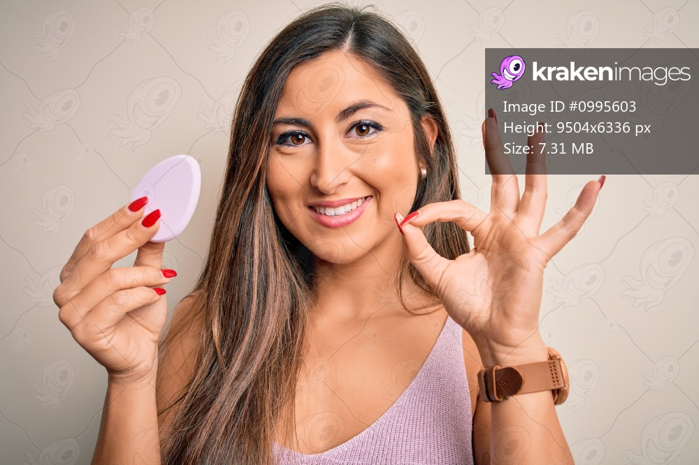 Young beautiful woman using beauty sponge with facial makeup over pink background doing ok sign with fingers, excellent symbol