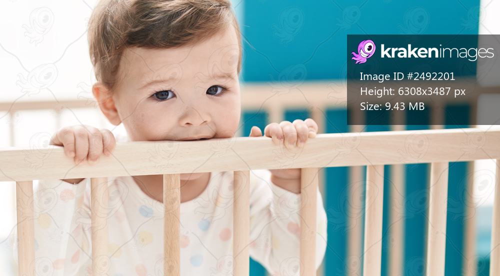 Adorable caucasian baby standing on cradle biting wooden balustrade at bedroom