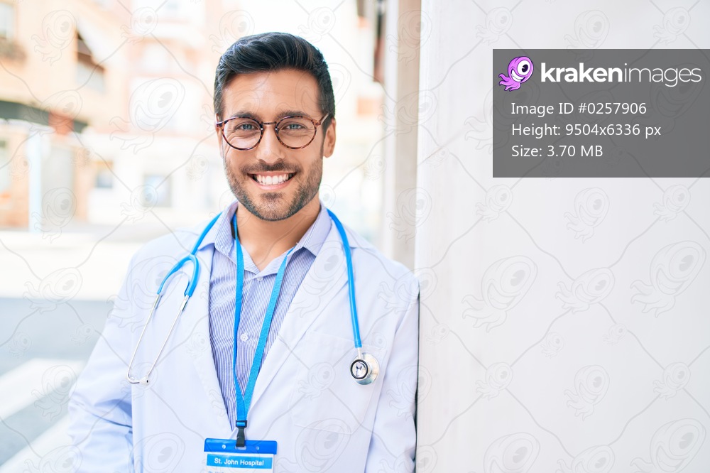 Young handsome hispanic doctor wearing uniform and stethoscope smiling happy. Standing with smile on face leaning on the wall at town street.