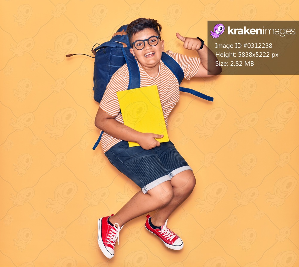 Adorable student boy wearing glasses and backpack smiling happy. Jumping with smile on face holding book doing ok sign with thumb up over isolated yellow background.