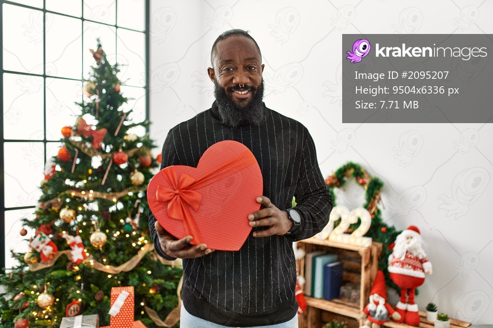 Young african american man holding heart box gift standing by christmas tree at home