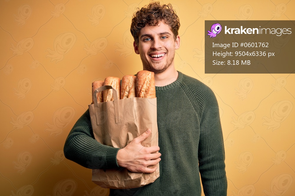 Young blond man with curly hair holding paper bag with bread over yellow background with a happy face standing and smiling with a confident smile showing teeth