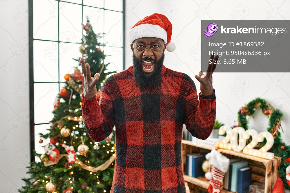 African american man wearing santa claus hat standing by christmas tree crazy and mad shouting and yelling with aggressive expression and arms raised. frustration concept. 