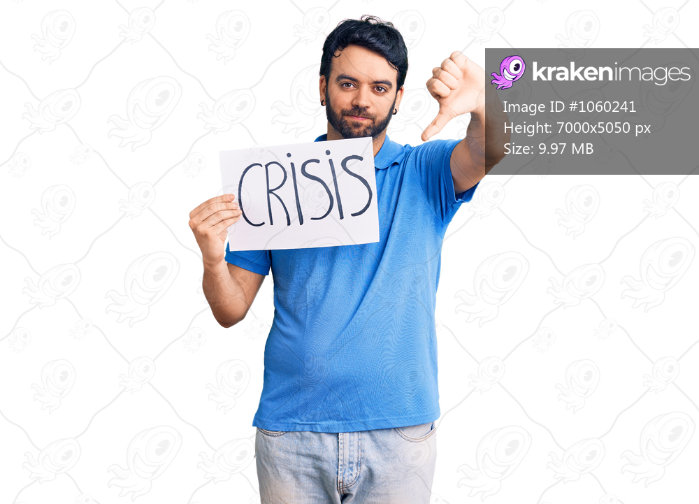 Young hispanic man holding crisis message paper with angry face, negative sign showing dislike with thumbs down, rejection concept 