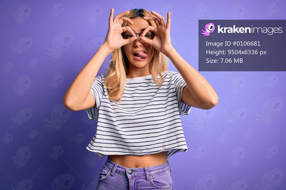 Young beautiful blonde woman wearing striped t-shirt and glasses over purple background doing ok gesture like binoculars sticking tongue out, eyes looking through fingers. Crazy expression.