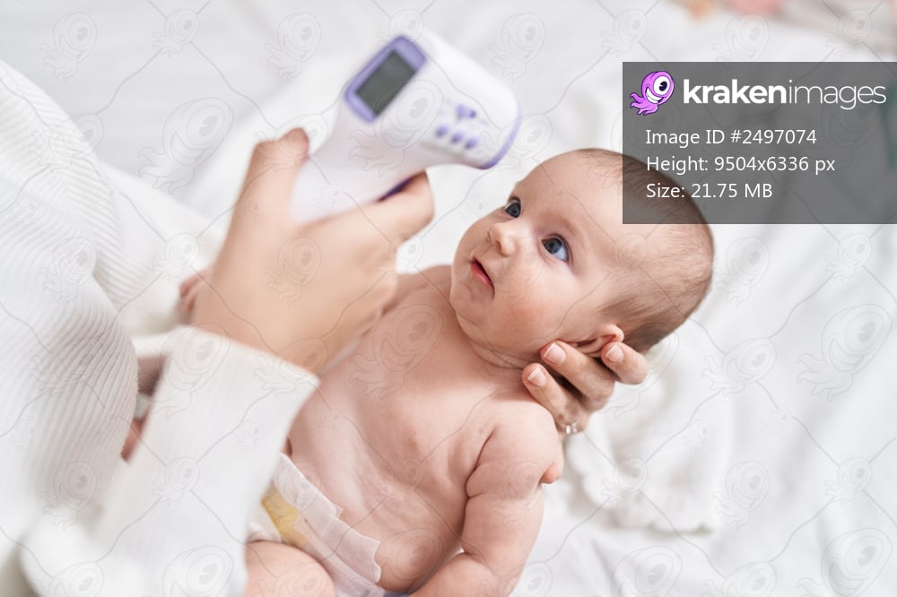 Adorable caucasian baby lying on bed measuring temperature with thermometer at bedroom