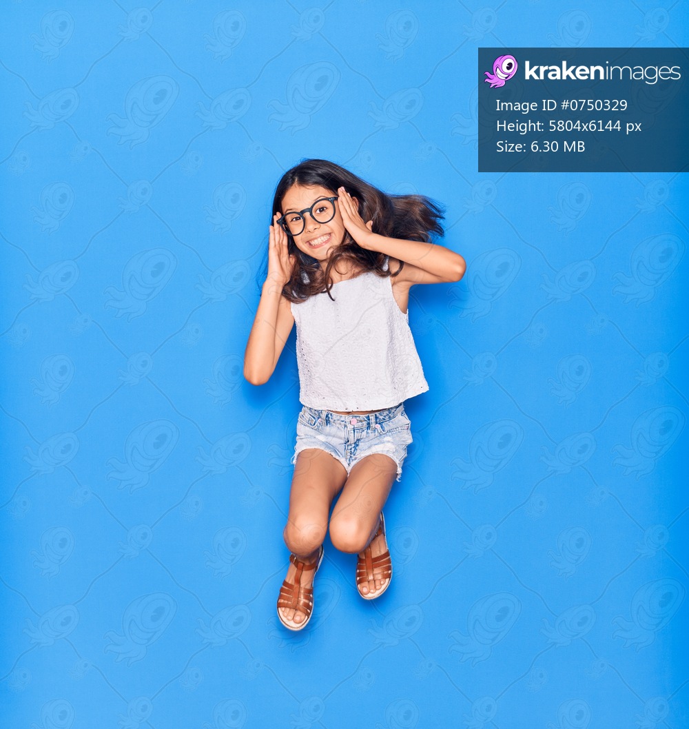 Adorable hispanic child girl wearing casual clothes and glasses smiling happy. Jumping with smile on face over isolated blue background