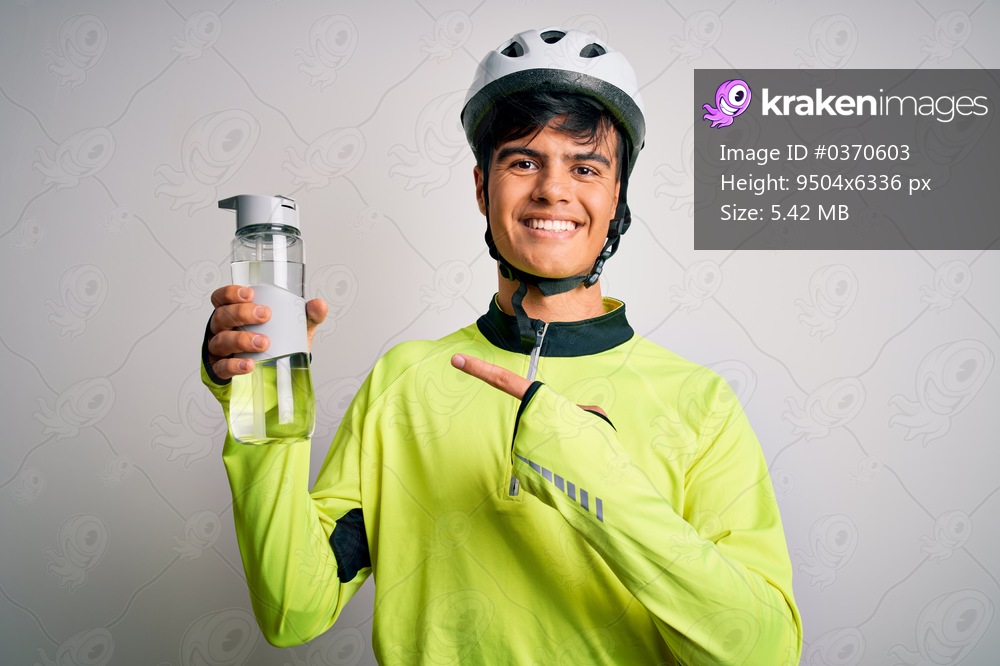 Young handsome cyclist man wearing security bike helmet drinking bottle of water very happy pointing with hand and finger