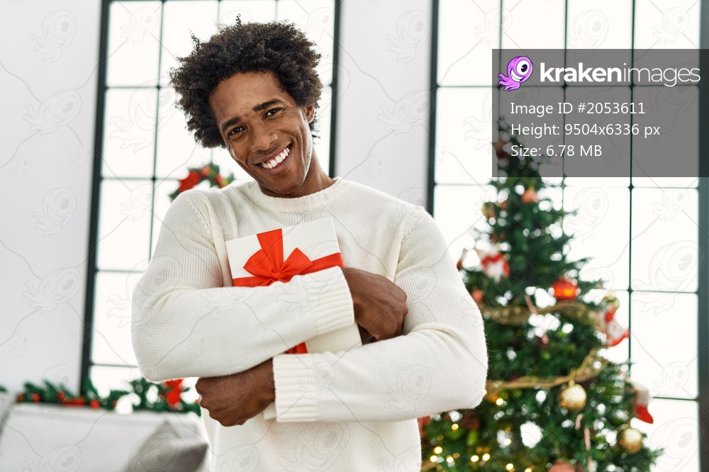 Young african american man holding gift standing by christmas tree at home.