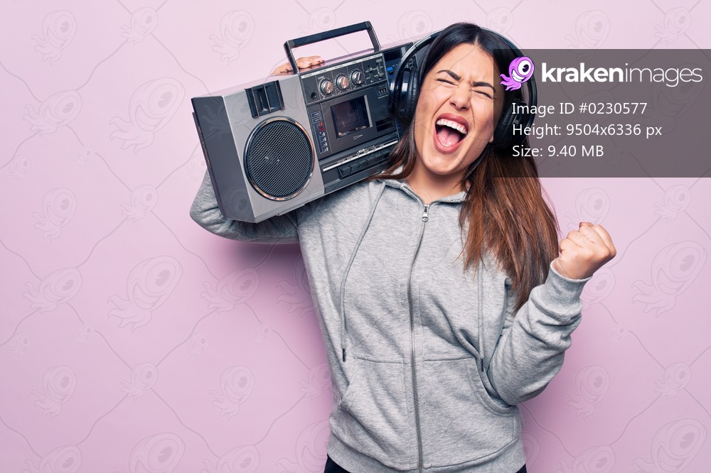 Young beautiful brunette woman listening to music using vintage boombox and headphones screaming proud, celebrating victory and success very excited with raised arm