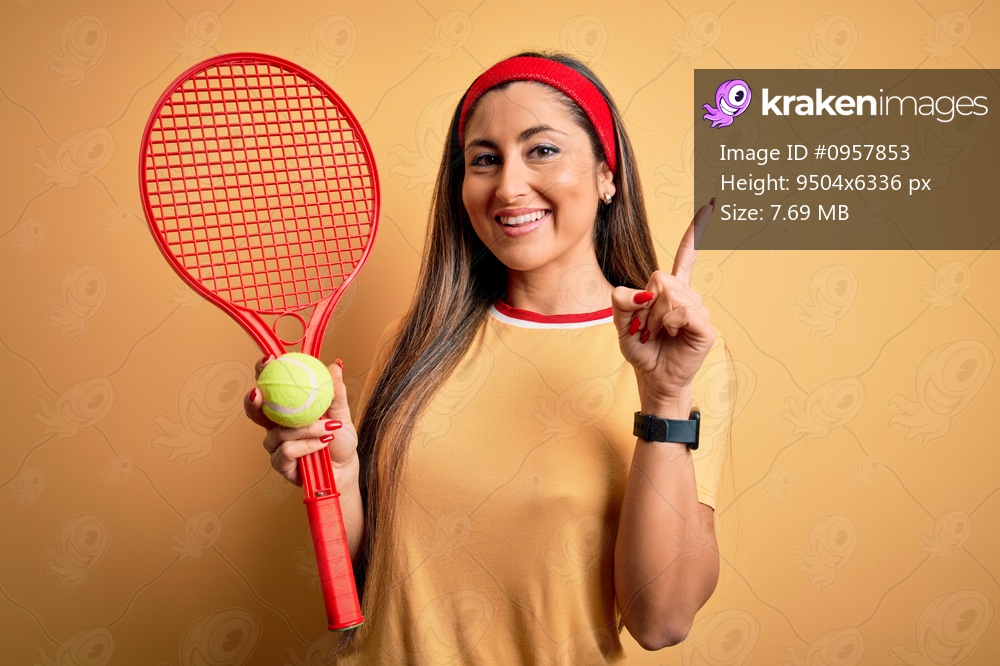 Beautiful brunette sportswoman playing tennis using racket and ball over yellow background surprised with an idea or question pointing finger with happy face, number one