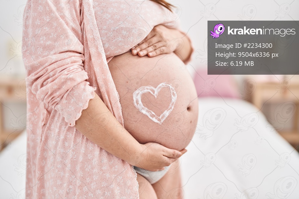 Young pregnant woman touching belly with heart lotion symbol standing at bedroom