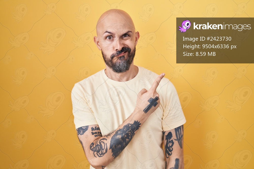Hispanic man with tattoos standing over yellow background pointing aside worried and nervous with forefinger, concerned and surprised expression 