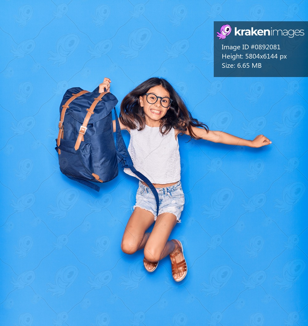 Adorable hispanic student child girl wearing glasses smiling happy. Jumping with smile on face holding backpack over isolated blue background