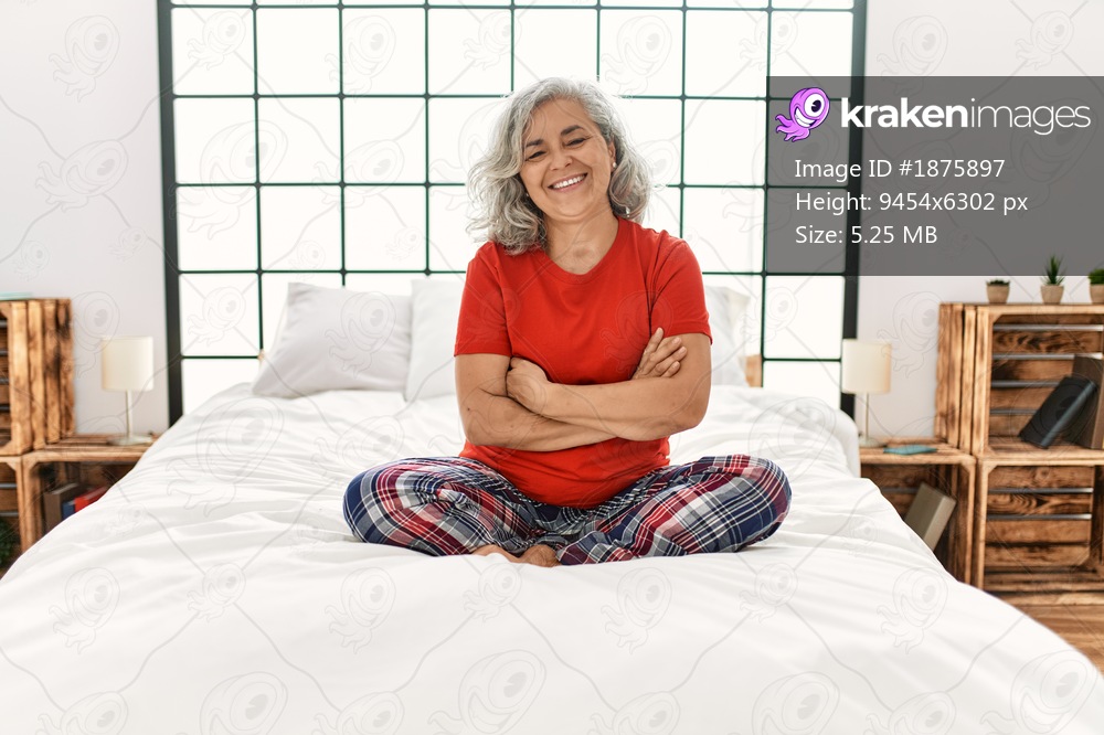 Middle age woman with grey hair sitting on the bed at home happy face smiling with crossed arms looking at the camera. positive person. 