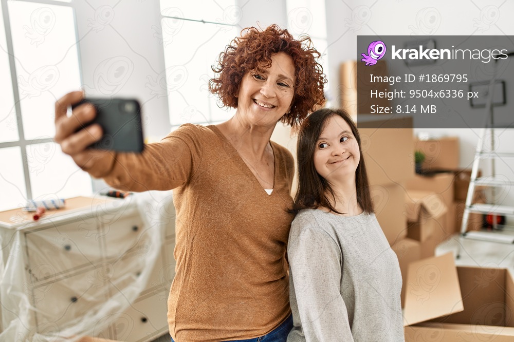 Mature mother and down syndrome daughter moving to a new home, standing by cardboard boxes taking a selfie picture