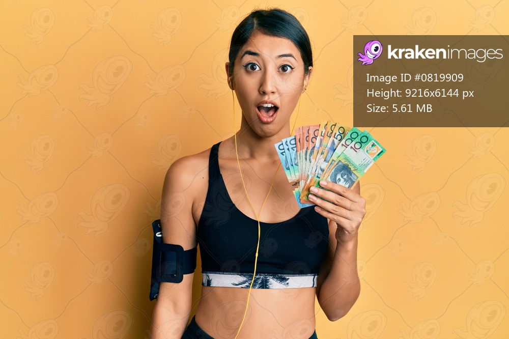 Beautiful asian young sport woman holding australian dollars scared and amazed with open mouth for surprise, disbelief face 