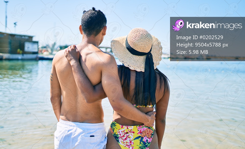 Young latin couple wearing swimwear hugging and   walking on back view at the beach.
