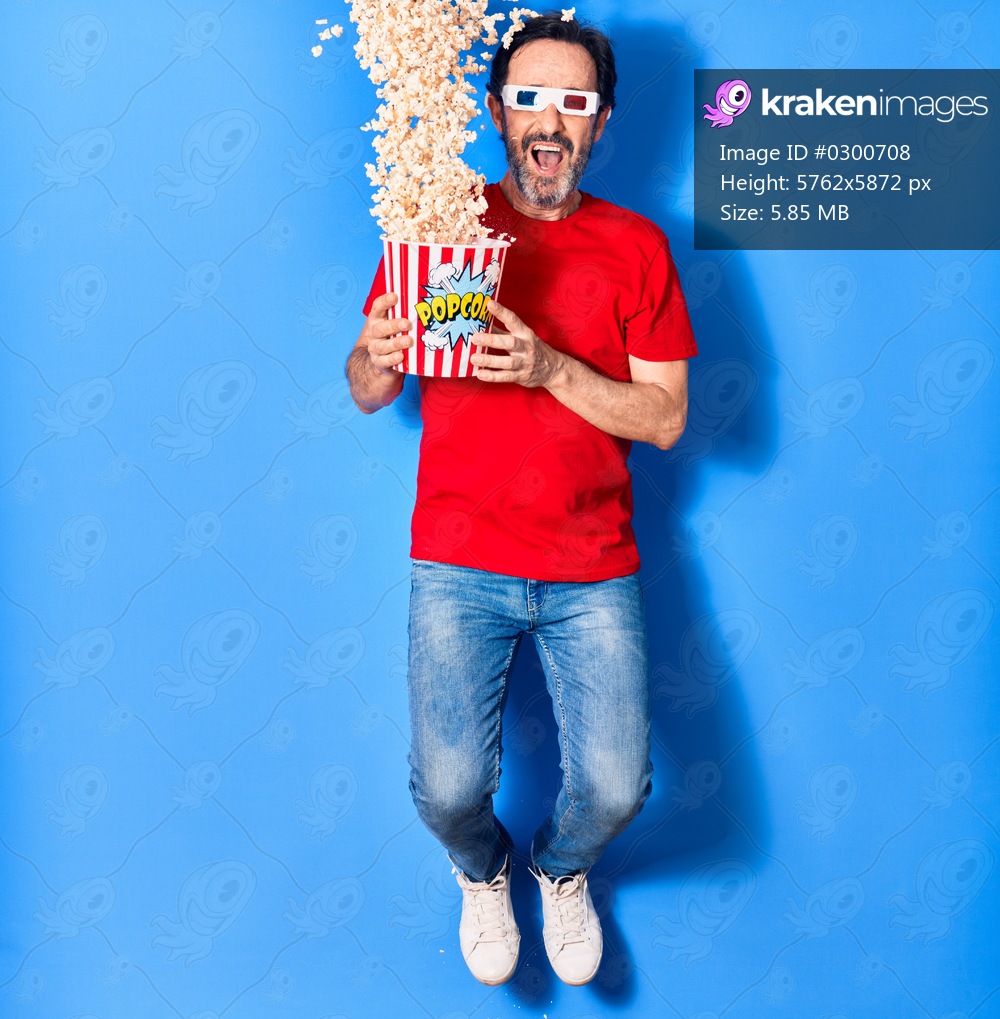 Middle age handsome hispanic man watching movie using 3d glasses similing happy. Holding bucket of popcorn jumping with smile on face over isolated blue background