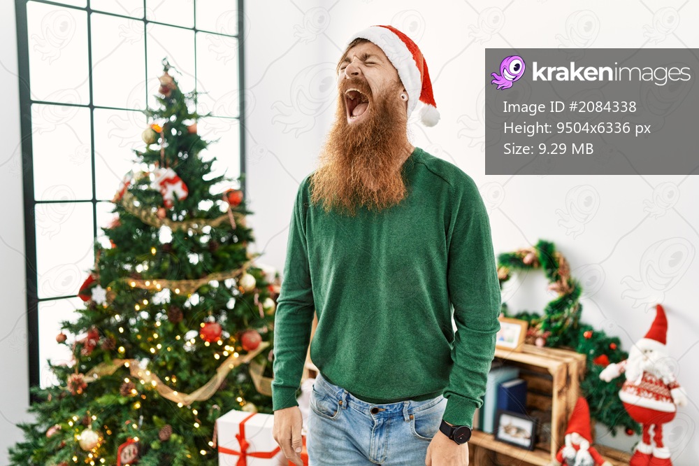 Redhead man with long beard wearing christmas hat by christmas tree angry and mad screaming frustrated and furious, shouting with anger. rage and aggressive concept. 